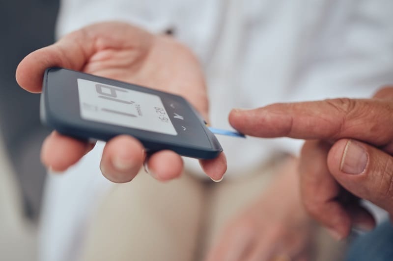 A close-up of a person's hand using a blood glucose meter to test blood sugar levels, with a healthcare professional assisting.