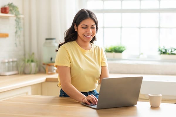 A woman in a yellow shirt sits at a kitchen table, smiling while using a laptop. A cup is placed beside her.